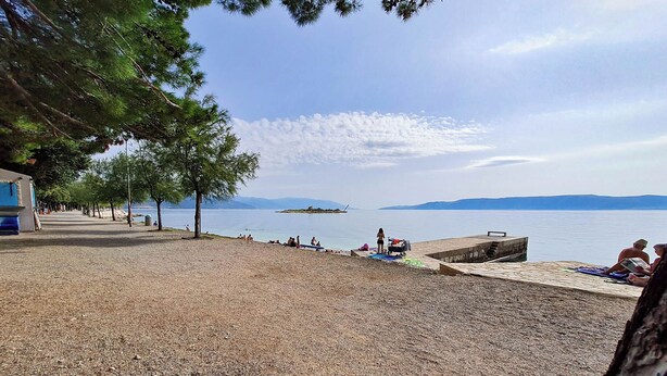 Una spiaggia con ciottoli, persone che prendono il sole e nuotano, una piccola isola all'orizzonte e un cielo sereno.