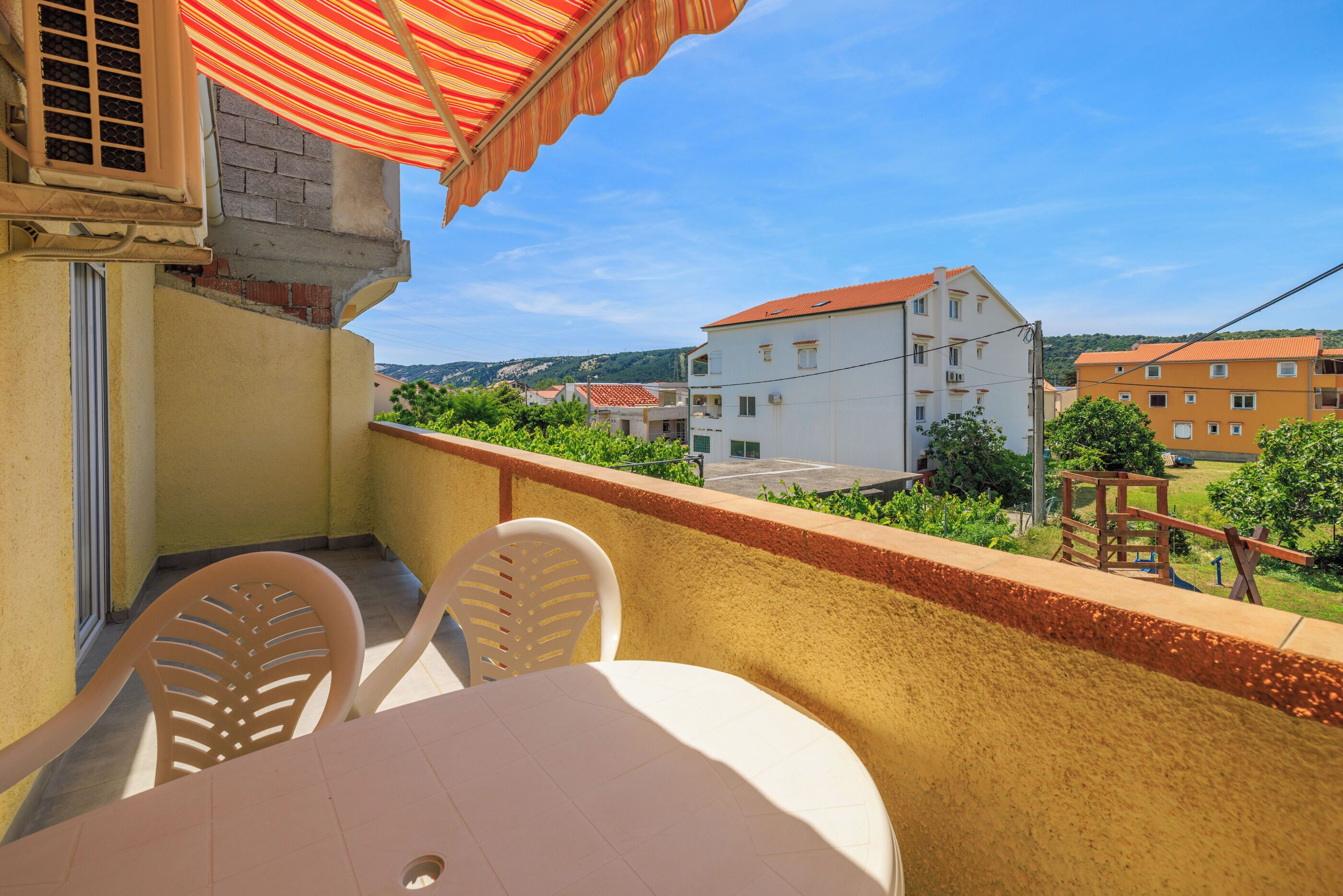 A balcony with a table and chairs, overlooking a street with buildings and greenery.