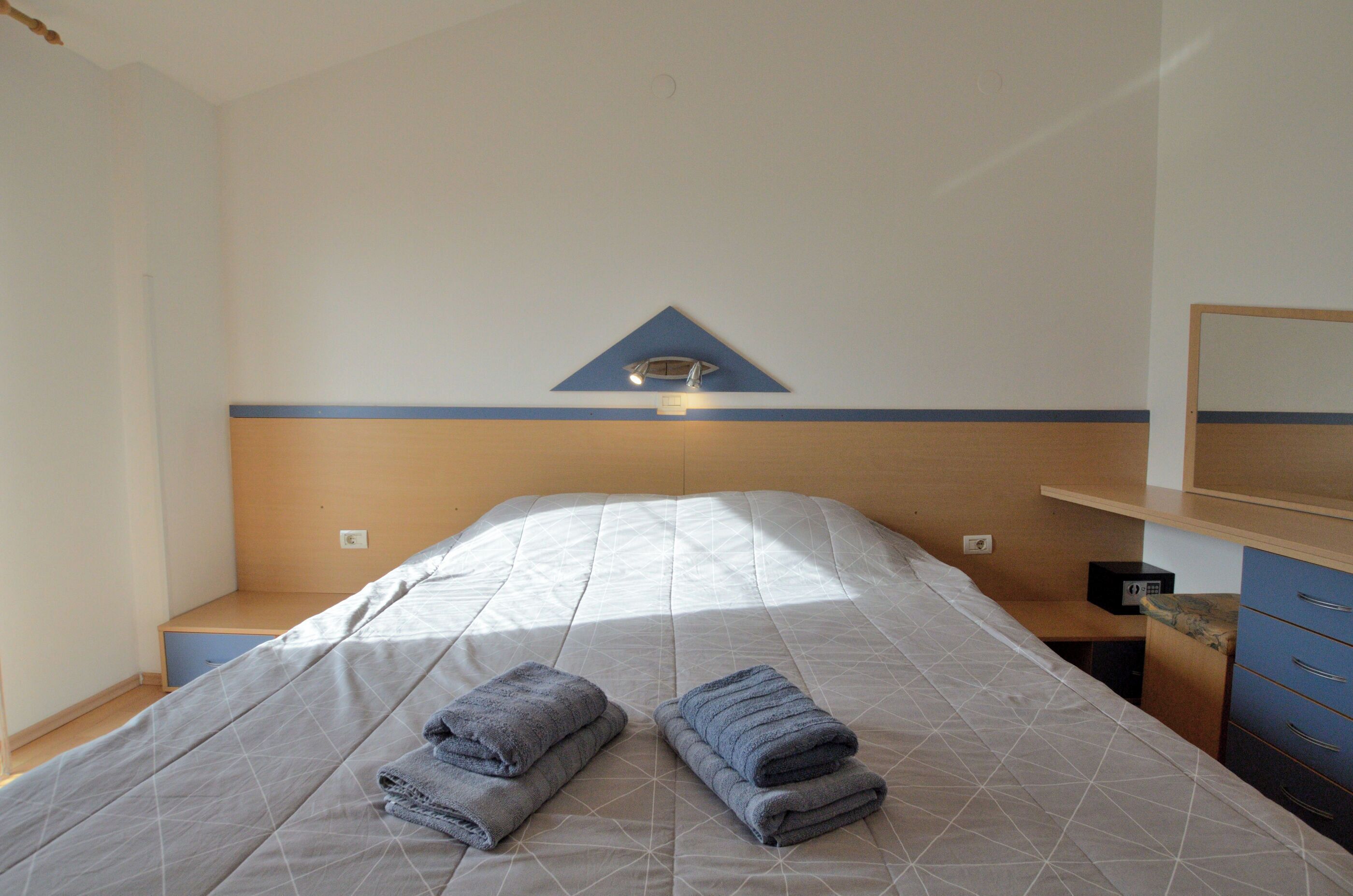 Una cama bien hecha con dos toallas dobladas, una mesita de noche y una lámpara de pared.