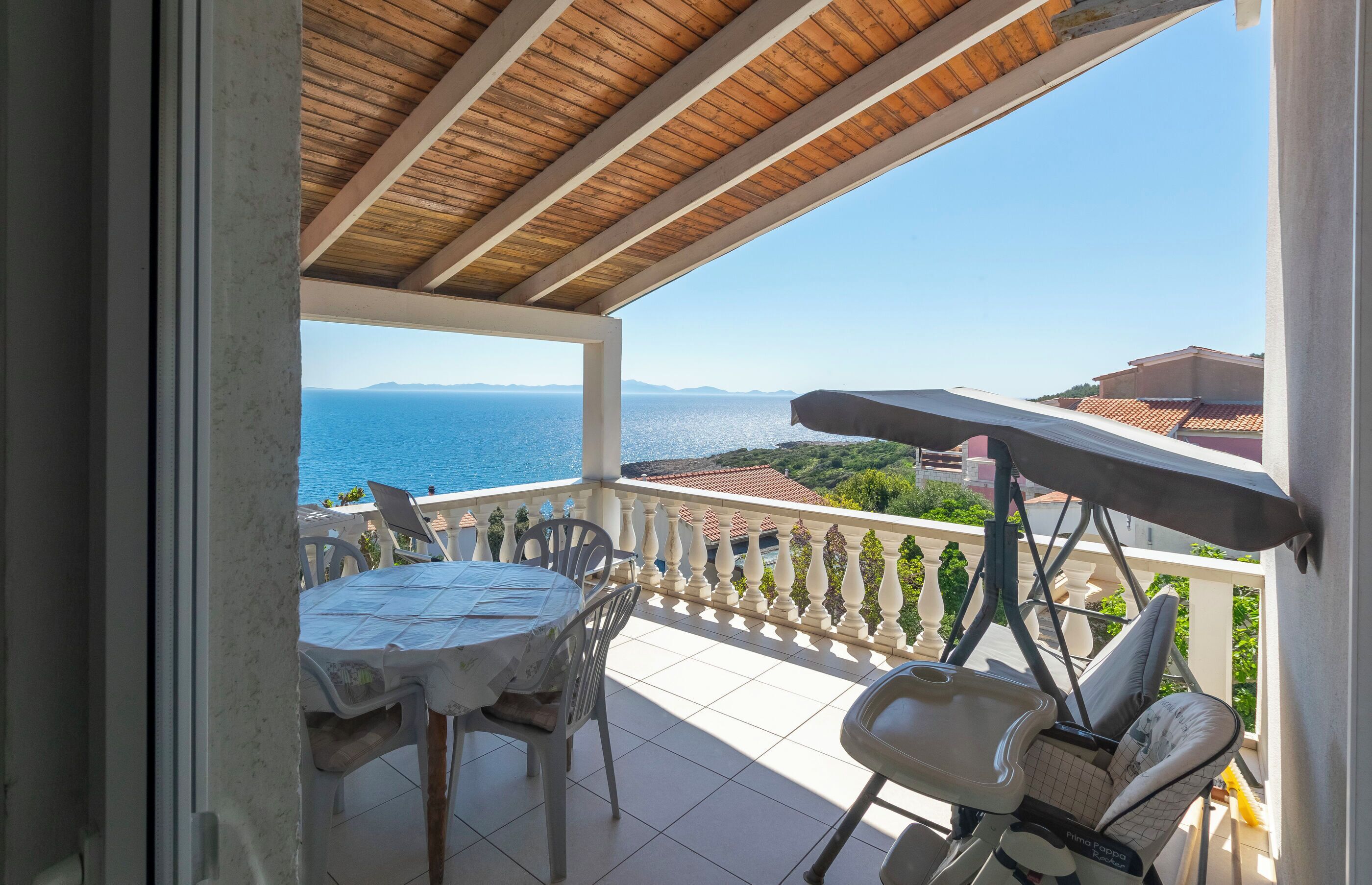 A balcony with a table and chairs, a baby stroller, and a view of the sea and distant hills.