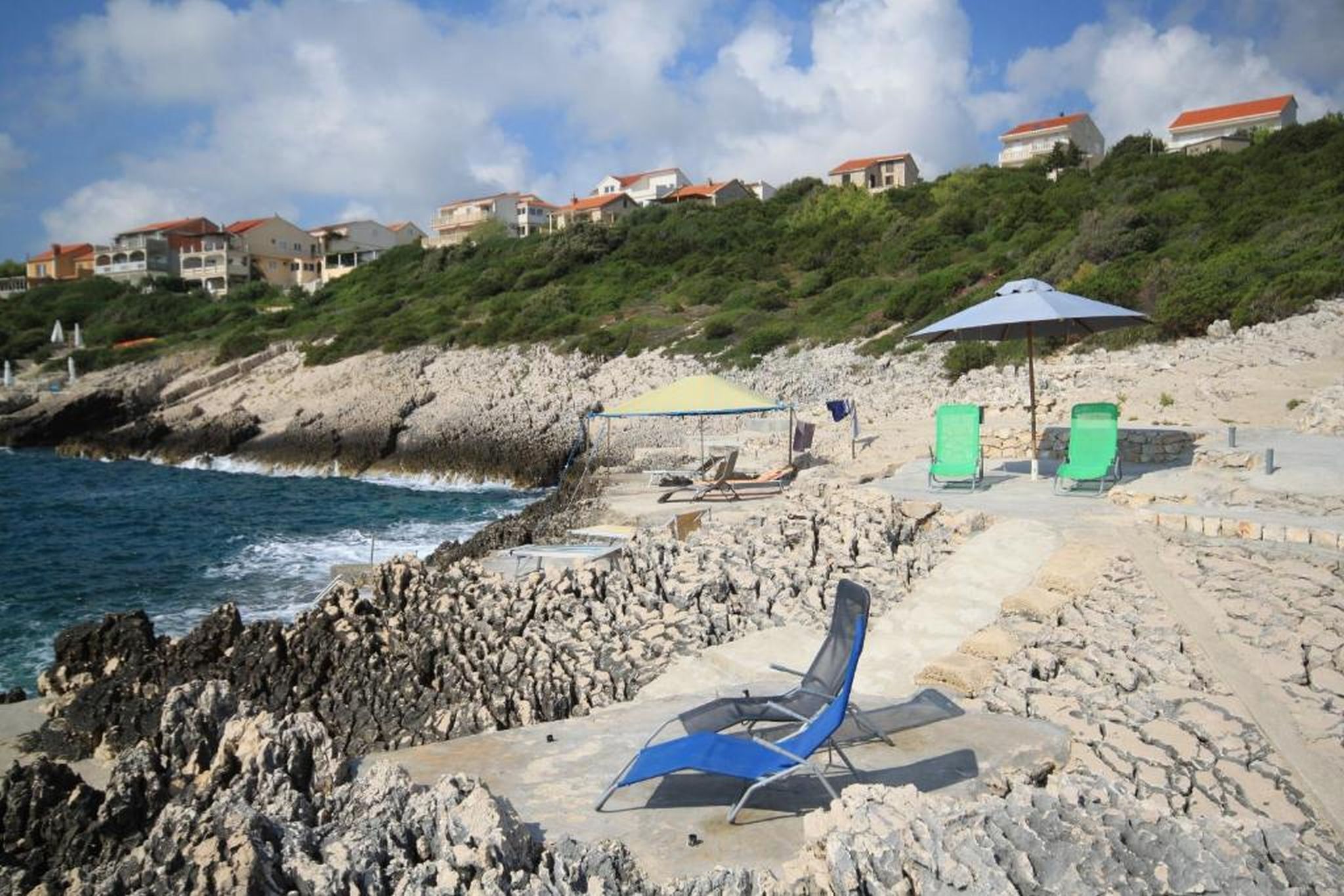 Uma praia rochosa com uma cadeira e um guarda-sol, uma linha costeira pedregosa e casas ao fundo.