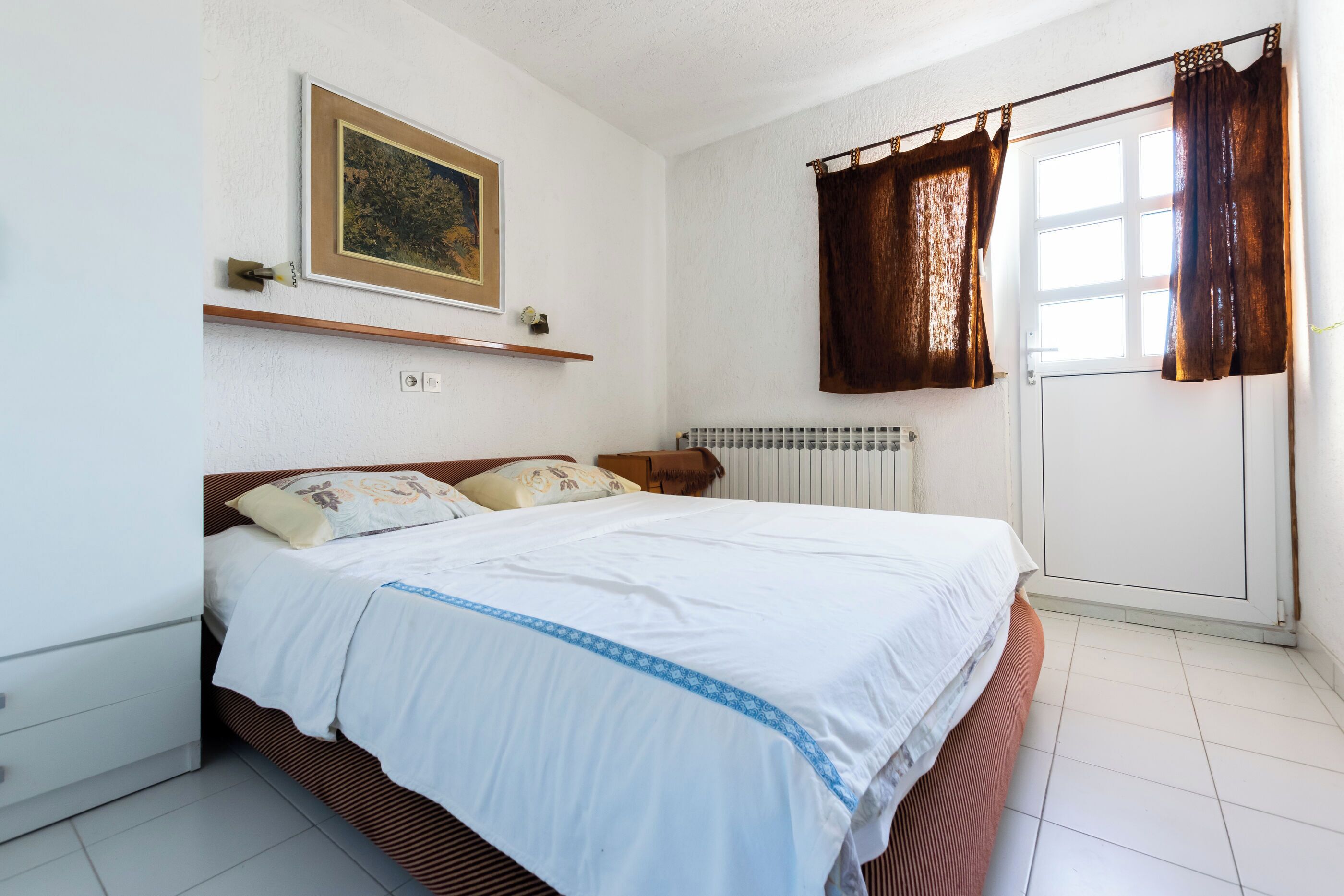 A bedroom with a bed, a radiator, a window with curtains, a shelf with a picture, and a door.