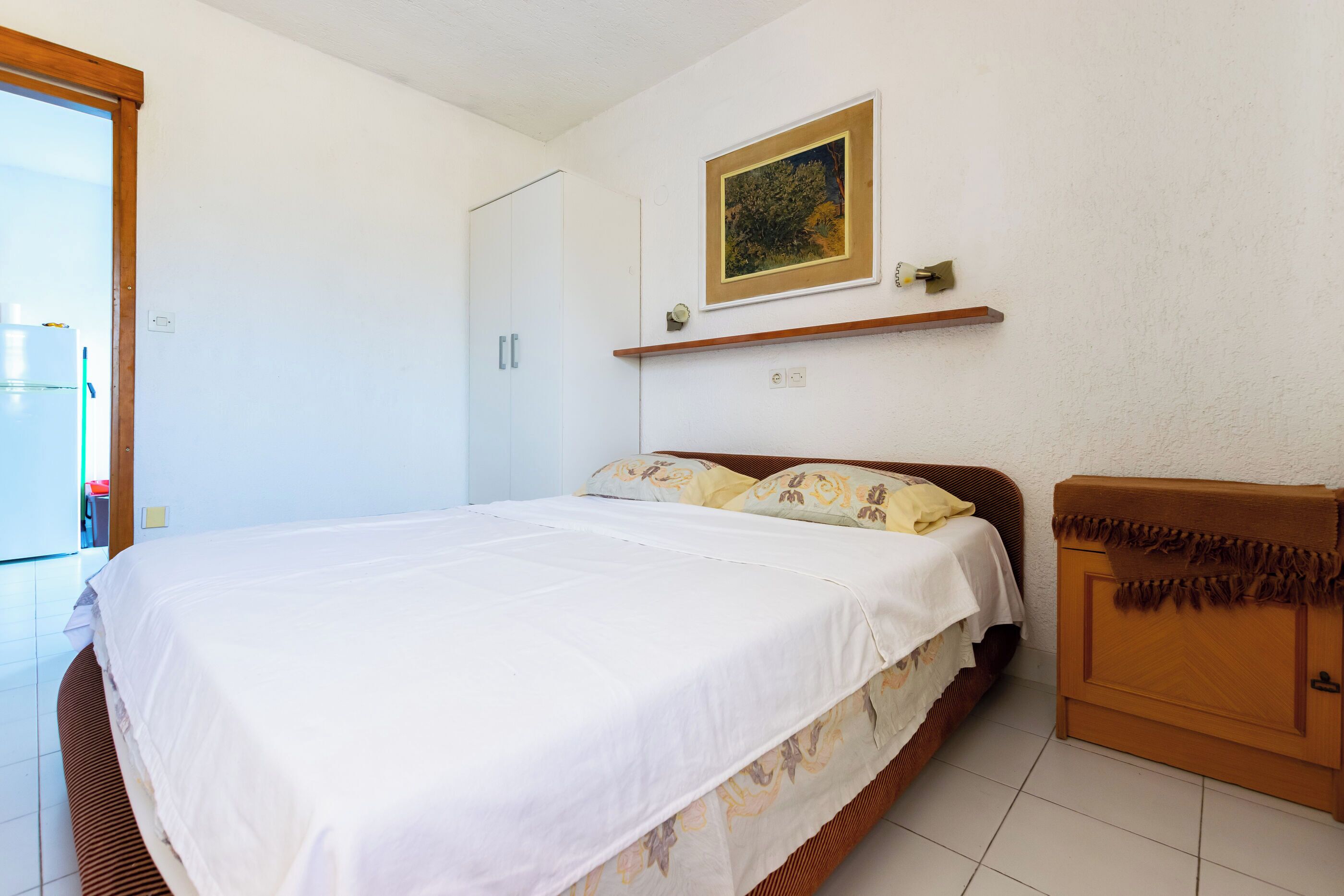 A bedroom with a bed, a wooden cabinet, a painting on the wall, and a door leading to another room.