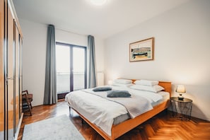 A hotel room with a wooden bed, a view of the sea through a large window, a painting on the wall, and a small round table with a lamp. - Apartment Peaceful Paradise (Bakar)