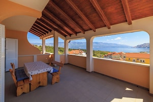 A covered balcony with a wooden table and chairs, a view of a lake and mountains, and a building with red-tiled roofs in the background.