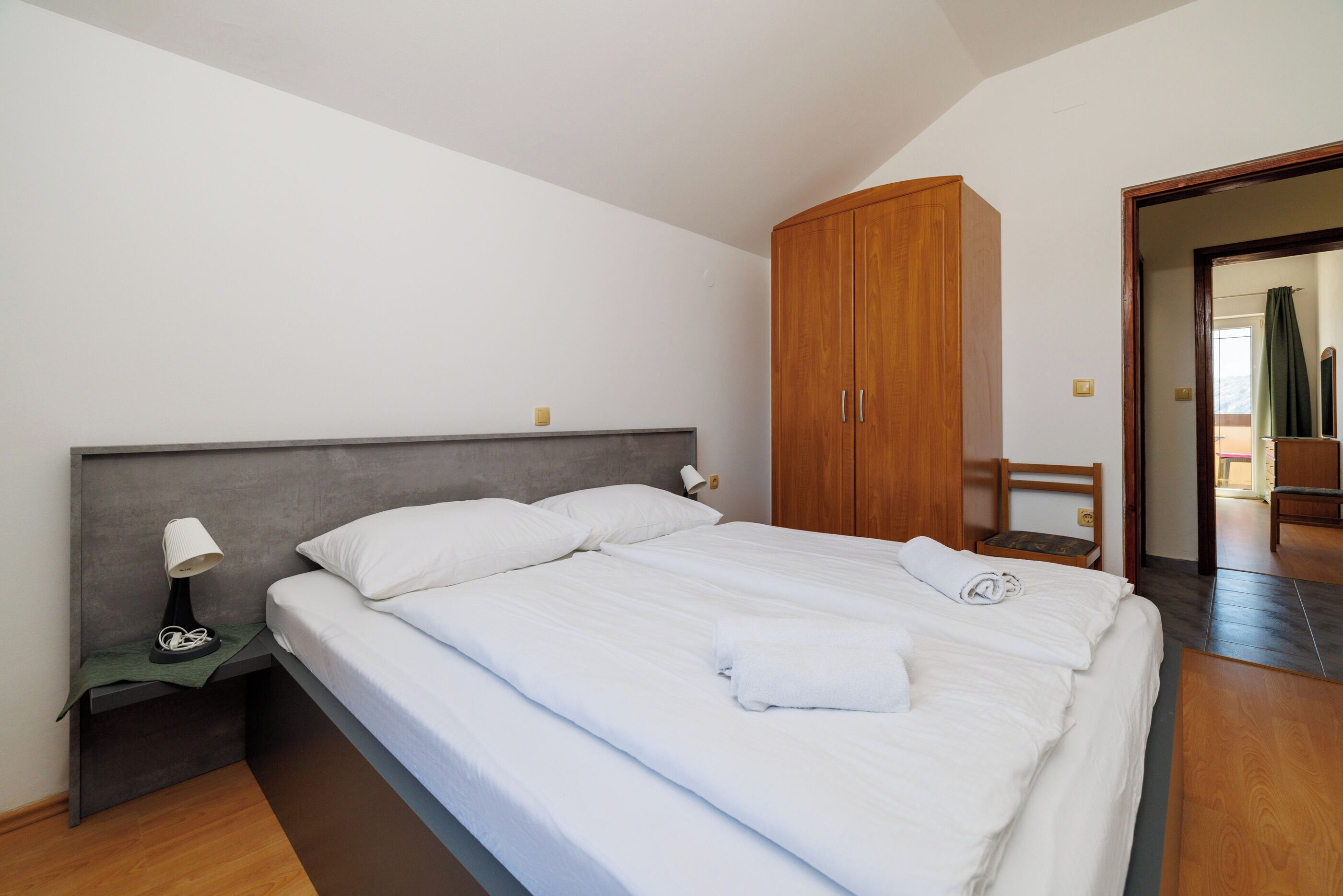 A neatly made bed with white linens, a wooden wardrobe, a bedside table with a lamp, and a chair in a modern room with wooden flooring and white walls.