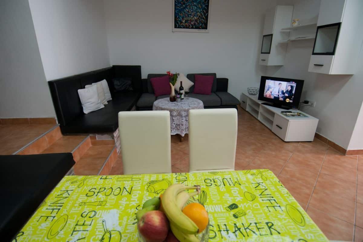 En stue med sofa, to stole, et spisebord med frugt, et fjernsyn og et lille bord med en vase og en vinflaske.