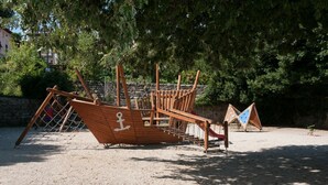 Een houten speelconstructie in de vorm van een schip in een zandrijk gebied, met bomen op de achtergrond.