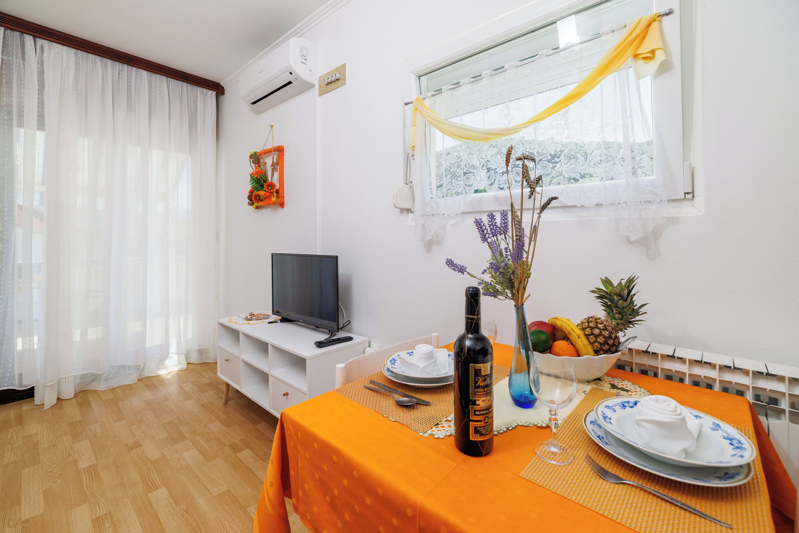 A dining area with a table set for two, a bottle of wine, a bowl of fruit, and a vase with flowers.