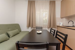 A dining area with a table, chairs, a green sofa, a window with curtains, and a kitchen area with cabinets and a sink. - Apartments Lemon (Vodice)