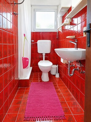 A bathroom with red tiles, a white toilet, a sink, and a pink rug. - Apartment Prcela 1 (Primosten)