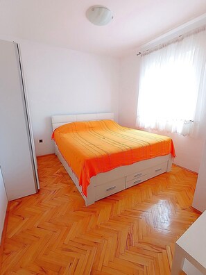 A bedroom with a bed, a wardrobe, a window with curtains, and a herringbone-patterned floor. - Apartment Prcela 1 (Primosten)