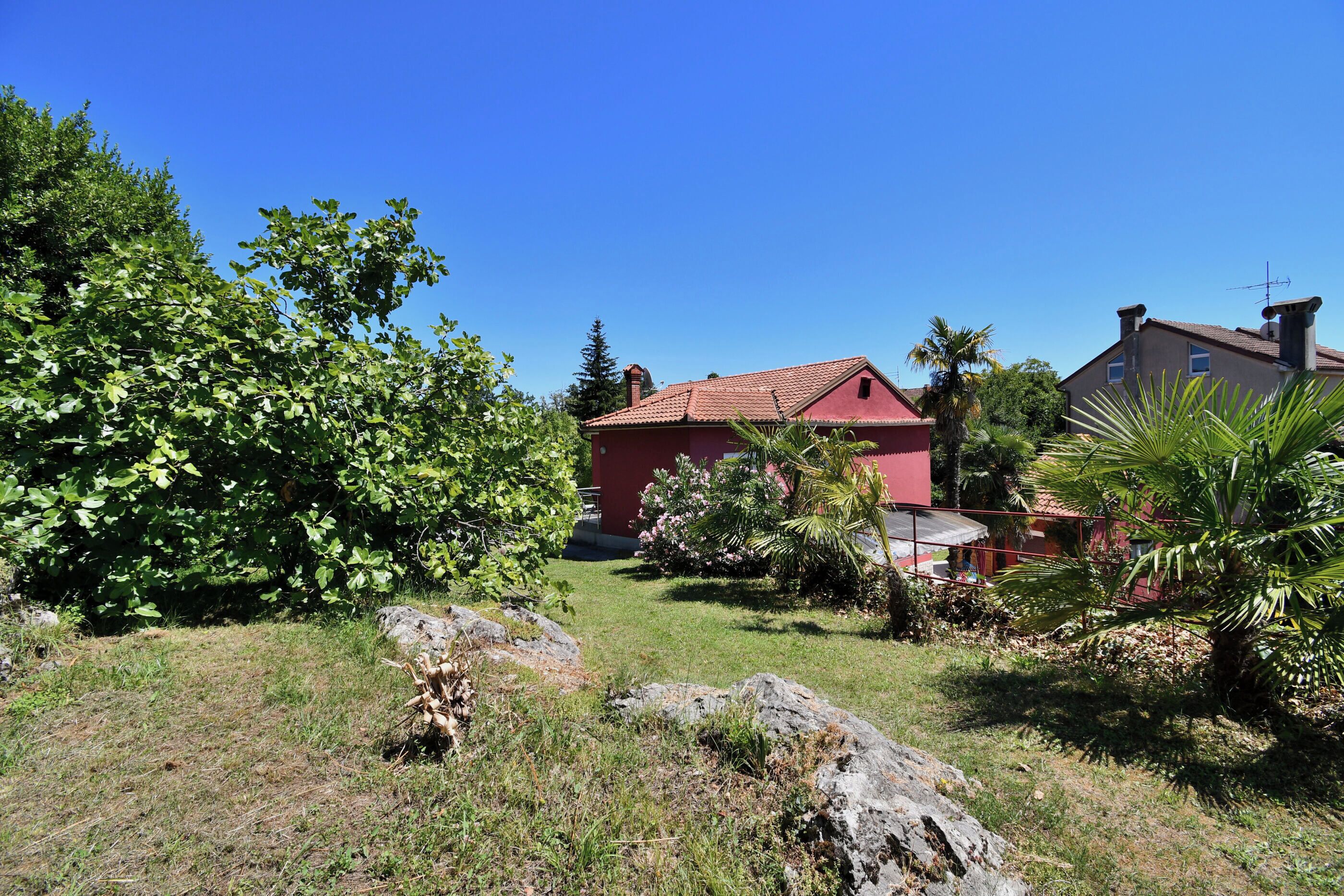 A red house with a tiled roof, surrounded by greenery and rocks.