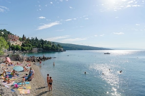 A beach with people sunbathing and swimming, a boat in the water, and buildings in the background. - House Iva i Ella (Opatija)