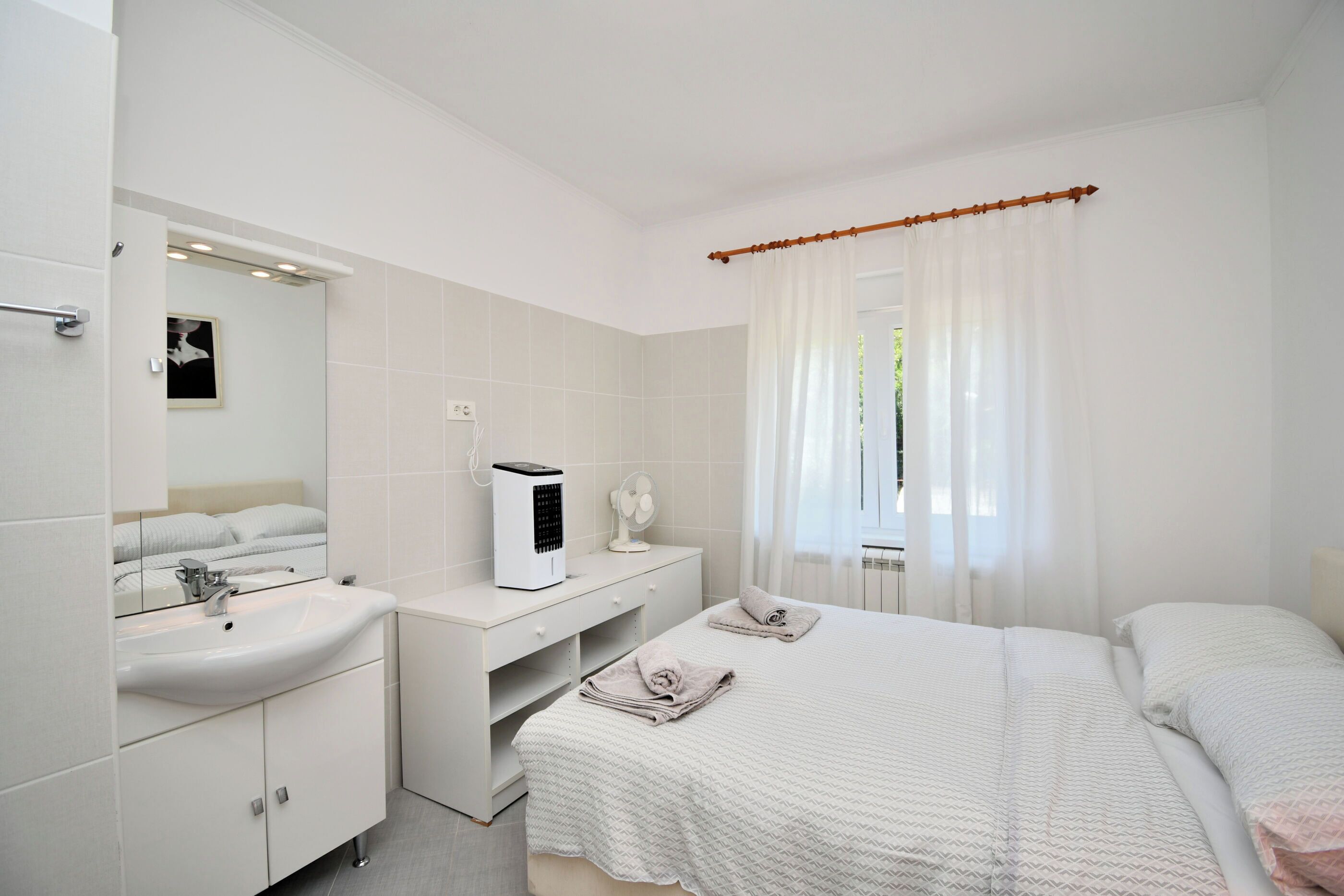 Une chambre moderne avec un grand lit, une table de chevet, un miroir, une commode blanche et une fenêtre agrémentée de rideaux légers.