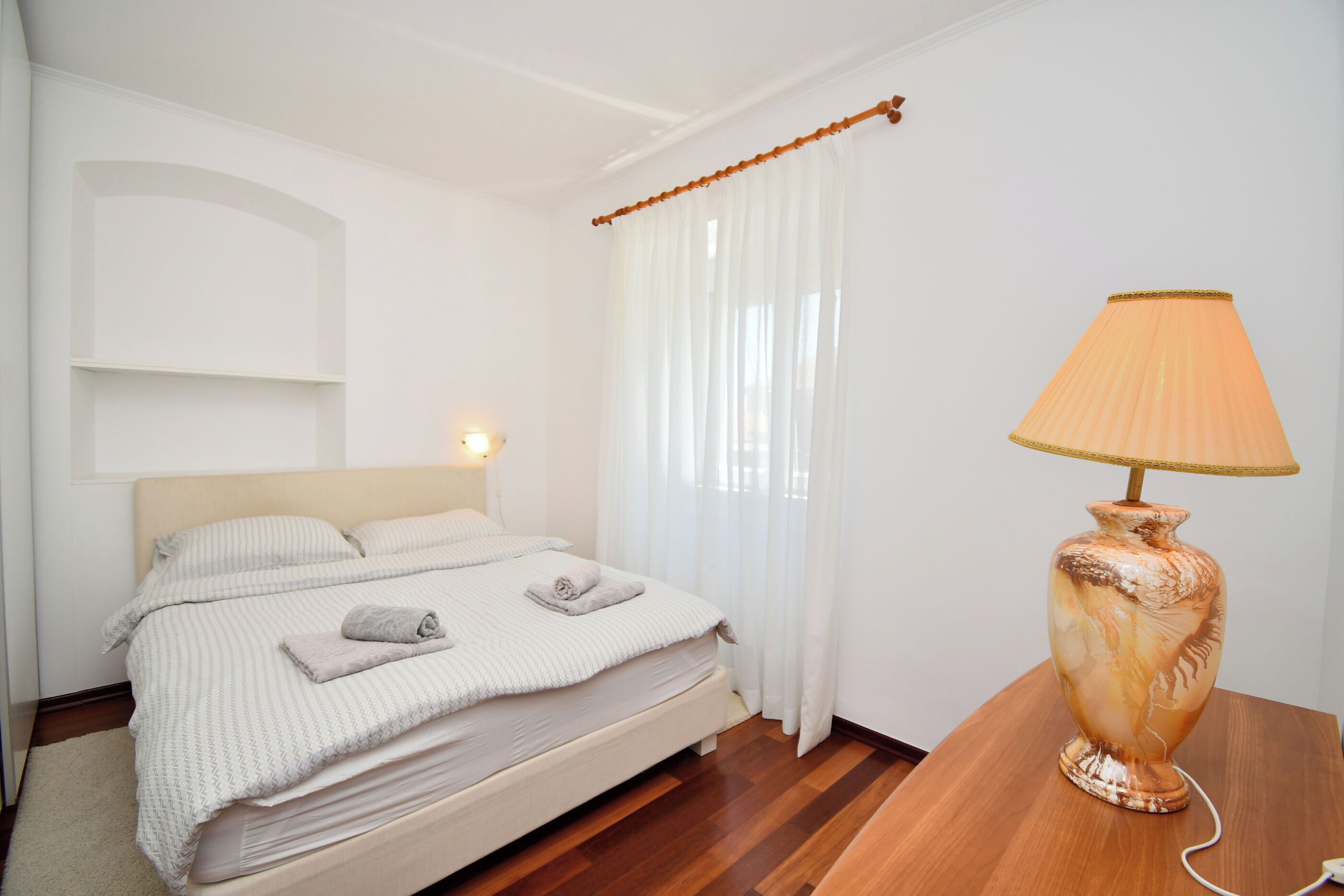 A neatly made bed with a white comforter and pillows, a bedside lamp, and a wooden nightstand.