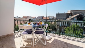 Un balcón con una sombrilla roja, sillas blancas, una mesa con bebidas y vistas a edificios.