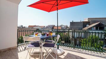 A balcony with a red umbrella, white chairs, a table with drinks, and a view of buildings.