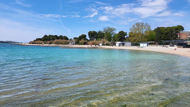 Una playa despejada con orilla de guijarros, una fila de cabañas de playa y árboles al fondo.