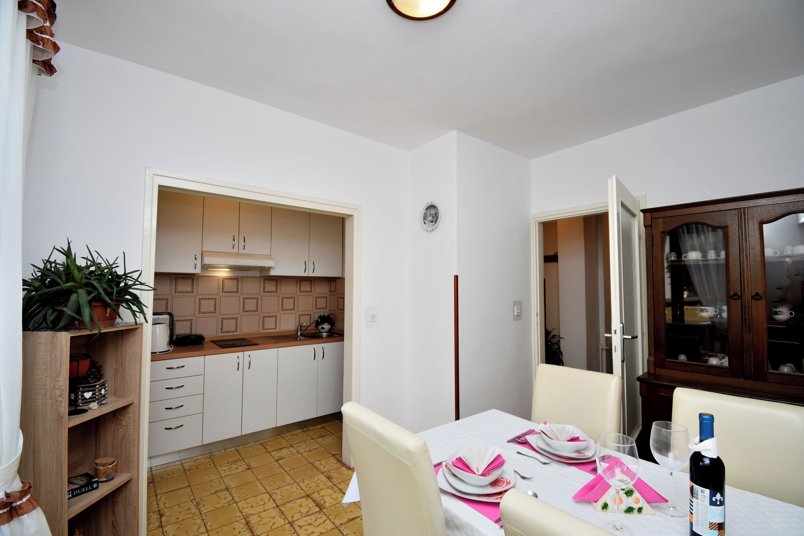 A dining area with a table set for two, a wine bottle, and a cabinet with dishes.
