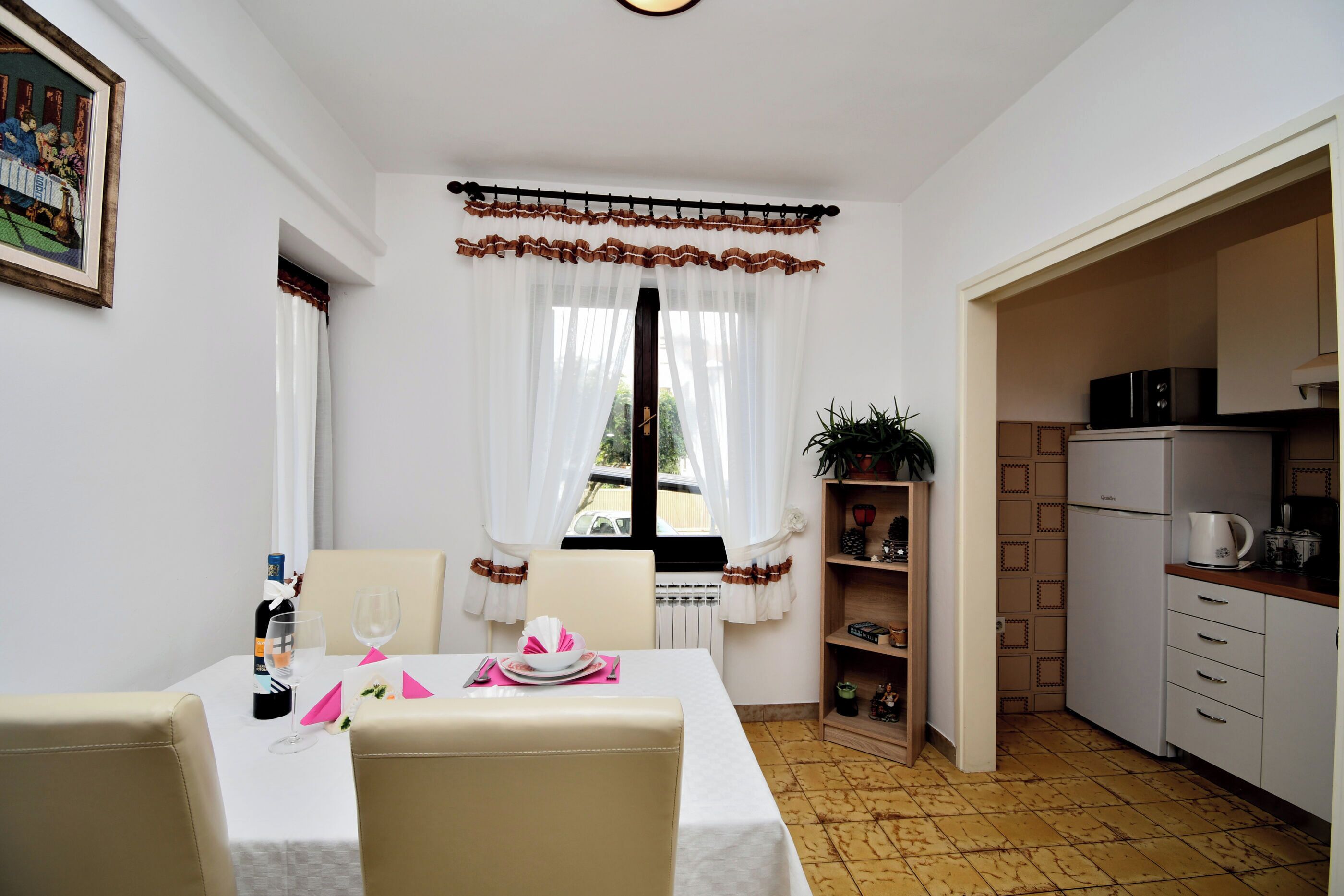A dining area with a table set for two, a kitchen with a refrigerator and microwave, and a window with curtains.