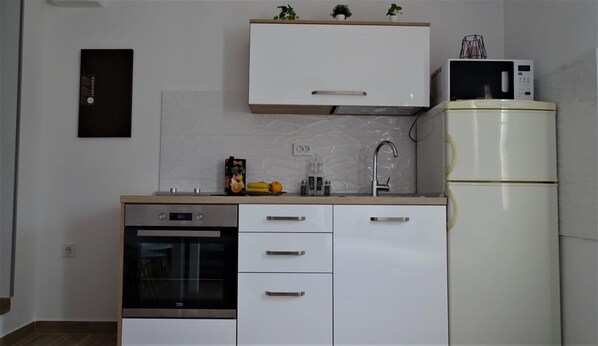 Eine kleine Küche mit Einbauofen, Mikrowelle und Kühlschrank. Es gibt Schränke mit Griffen, eine Spüle mit Wasserhahn und eine Arbeitsplatte mit einigen Gegenständen wie einem Buch und einer Obstschale.
