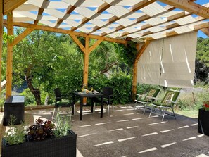 Ein überdachter Patio-Bereich mit Tisch und Stühlen, umgeben von Grünpflanzen und einer Pergola.