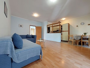 A living room with a sofa, a dining table with chairs, a kitchen area with a refrigerator, and a wooden floor. - Apartments Baburic (Krk)