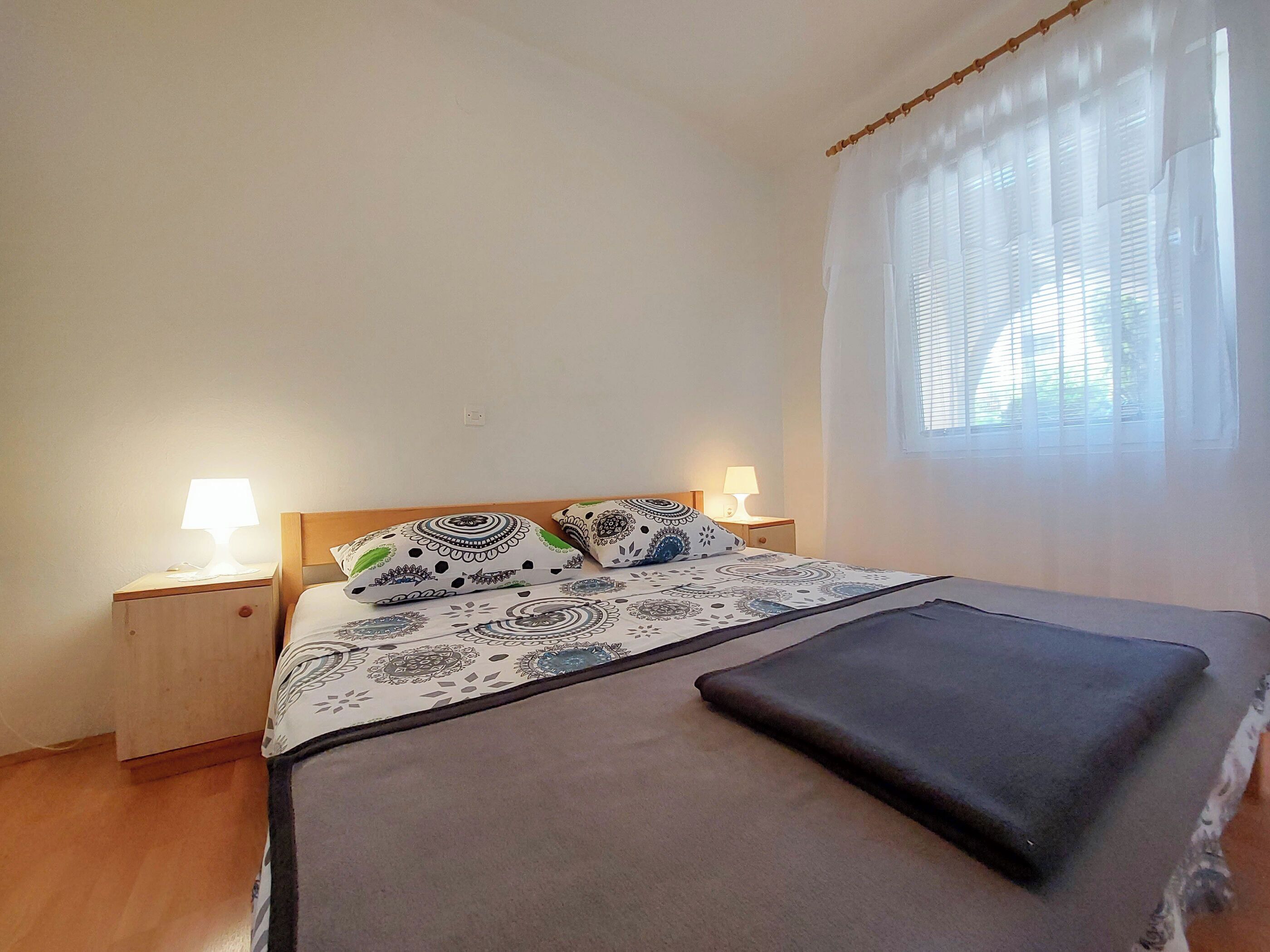 A bedroom with a bed, two bedside tables, a window with blinds, and a bedside lamp on each table.