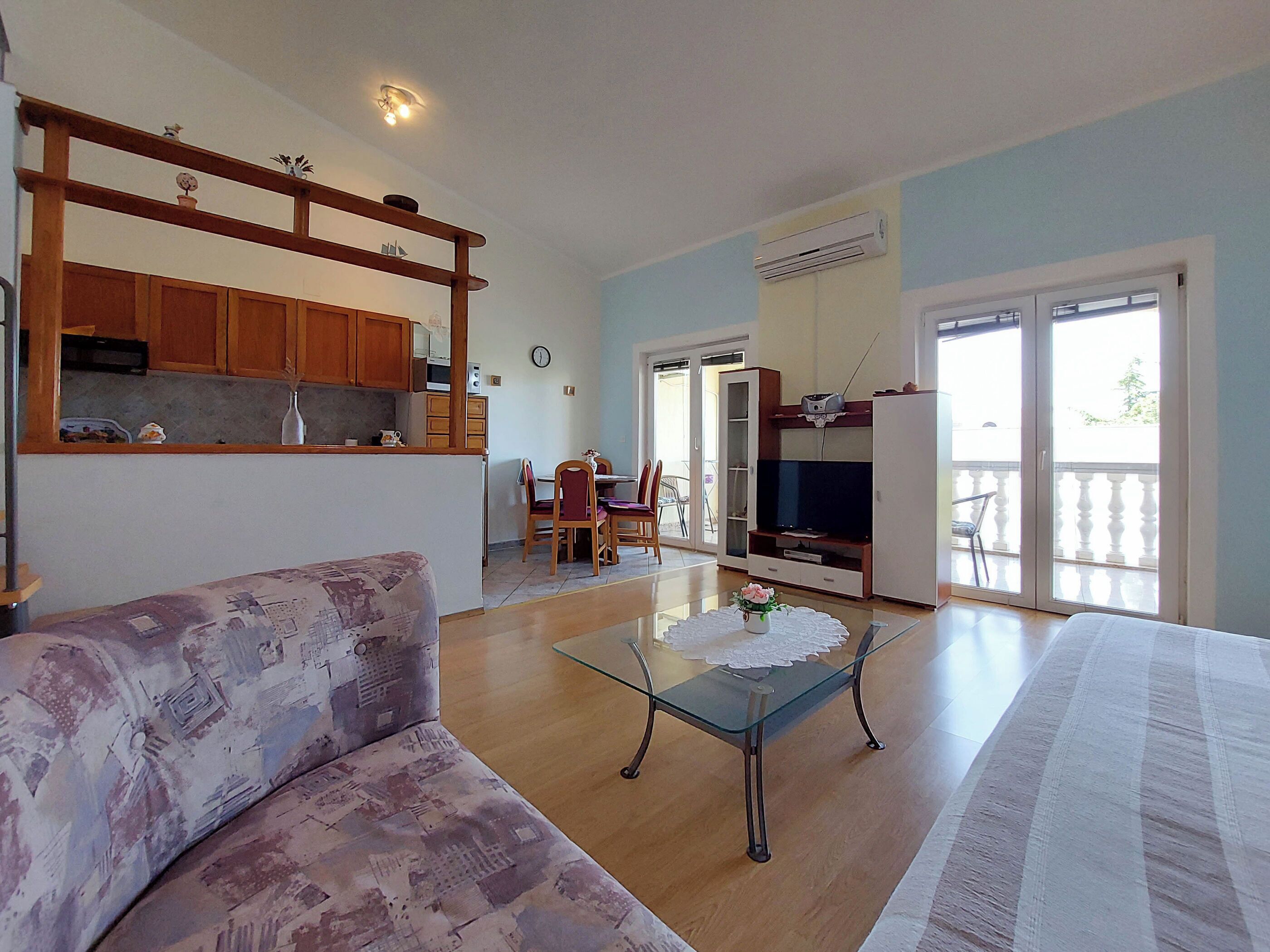 A living room with a sofa, a glass coffee table, and a television. There is a kitchen area with wooden cabinets and a dining area with chairs and a table.