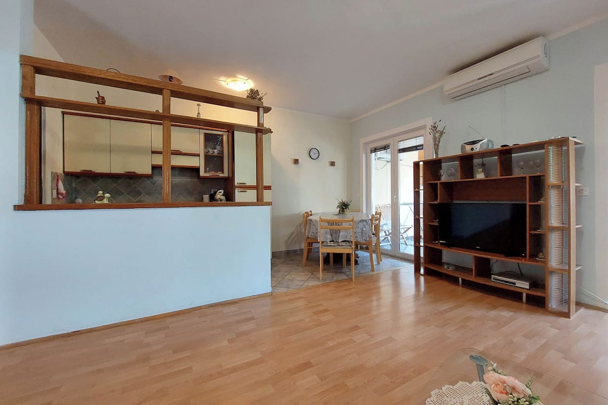 Un soggiorno con pavimento in legno, una zona pranzo con tavolo e sedie, un grande specchio e un mobile TV con televisore a schermo piatto.