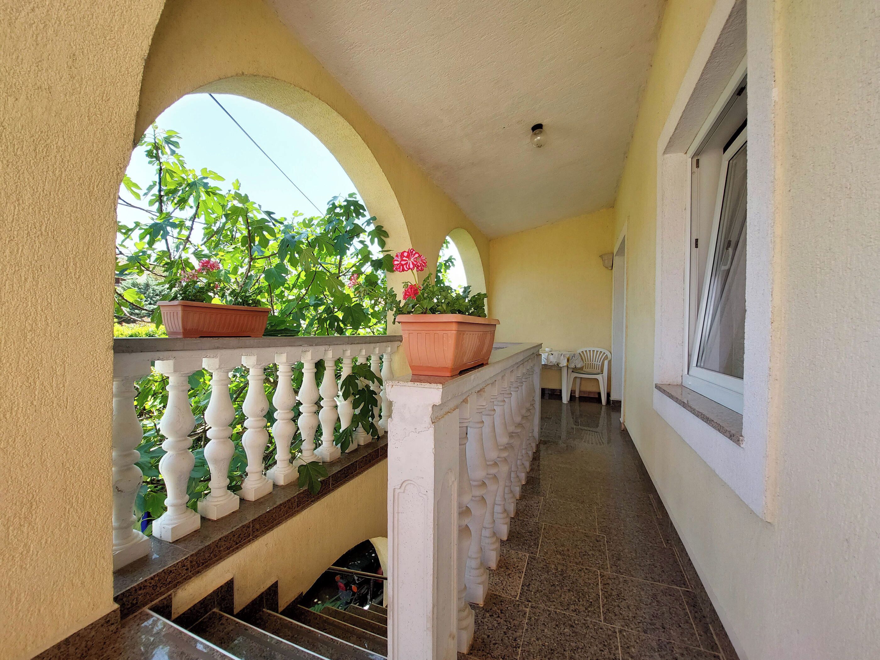 A covered balcony with potted plants, a white railing, and a window on the right.
