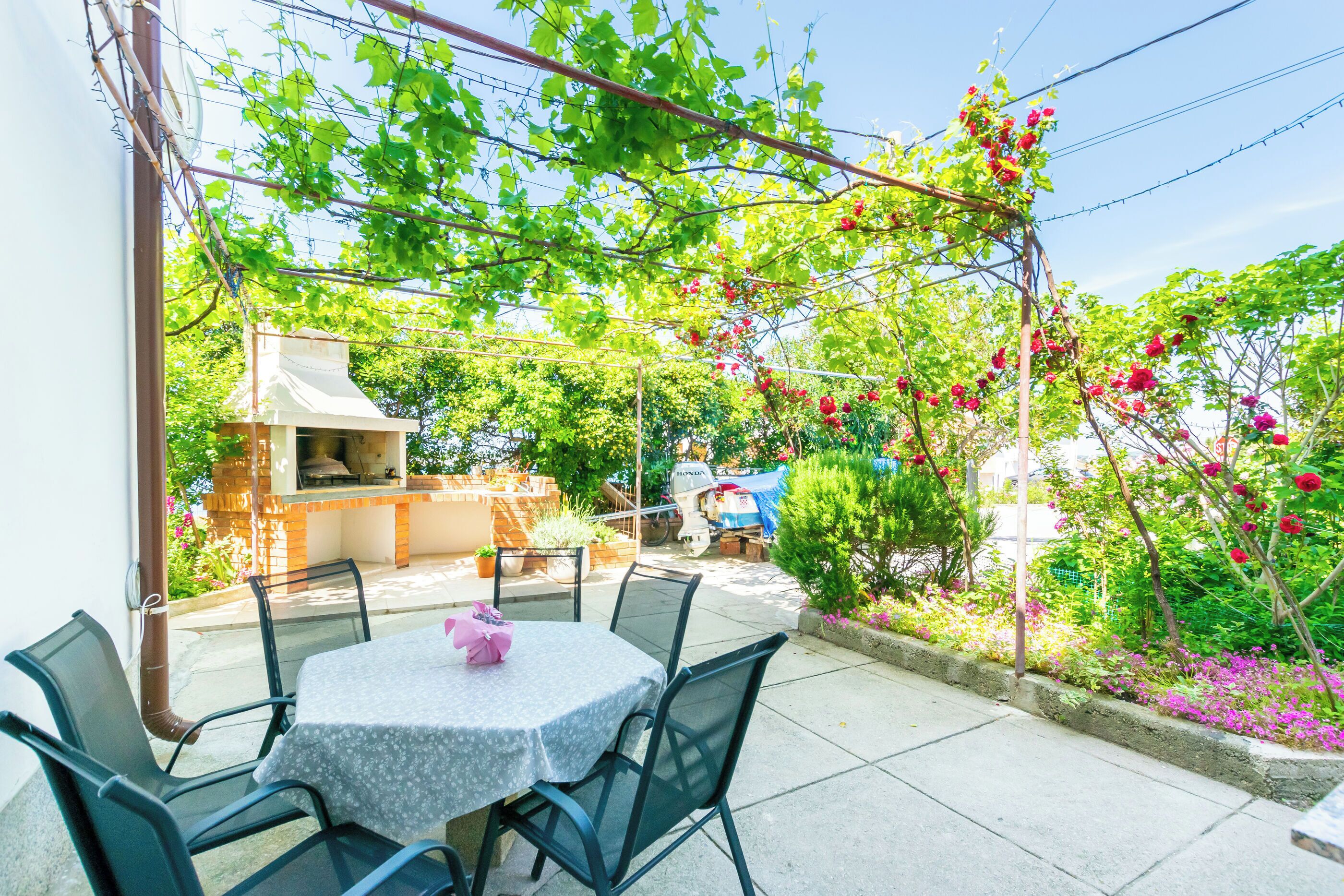 An outdoor patio with a round table and chairs, surrounded by lush greenery and flowering plants.