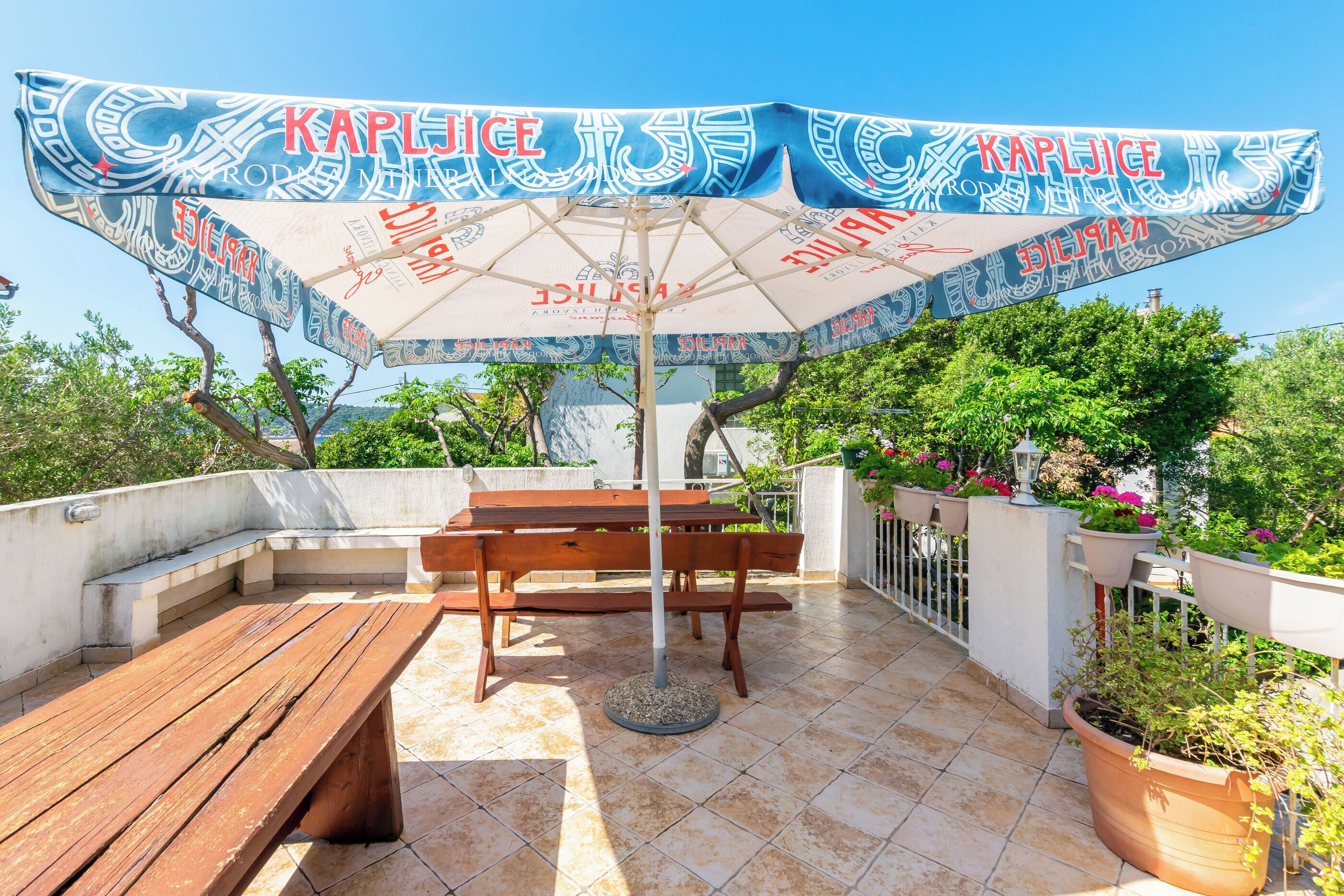 A patio with a wooden table and benches, an umbrella with 'Kaplice' branding, and potted plants.