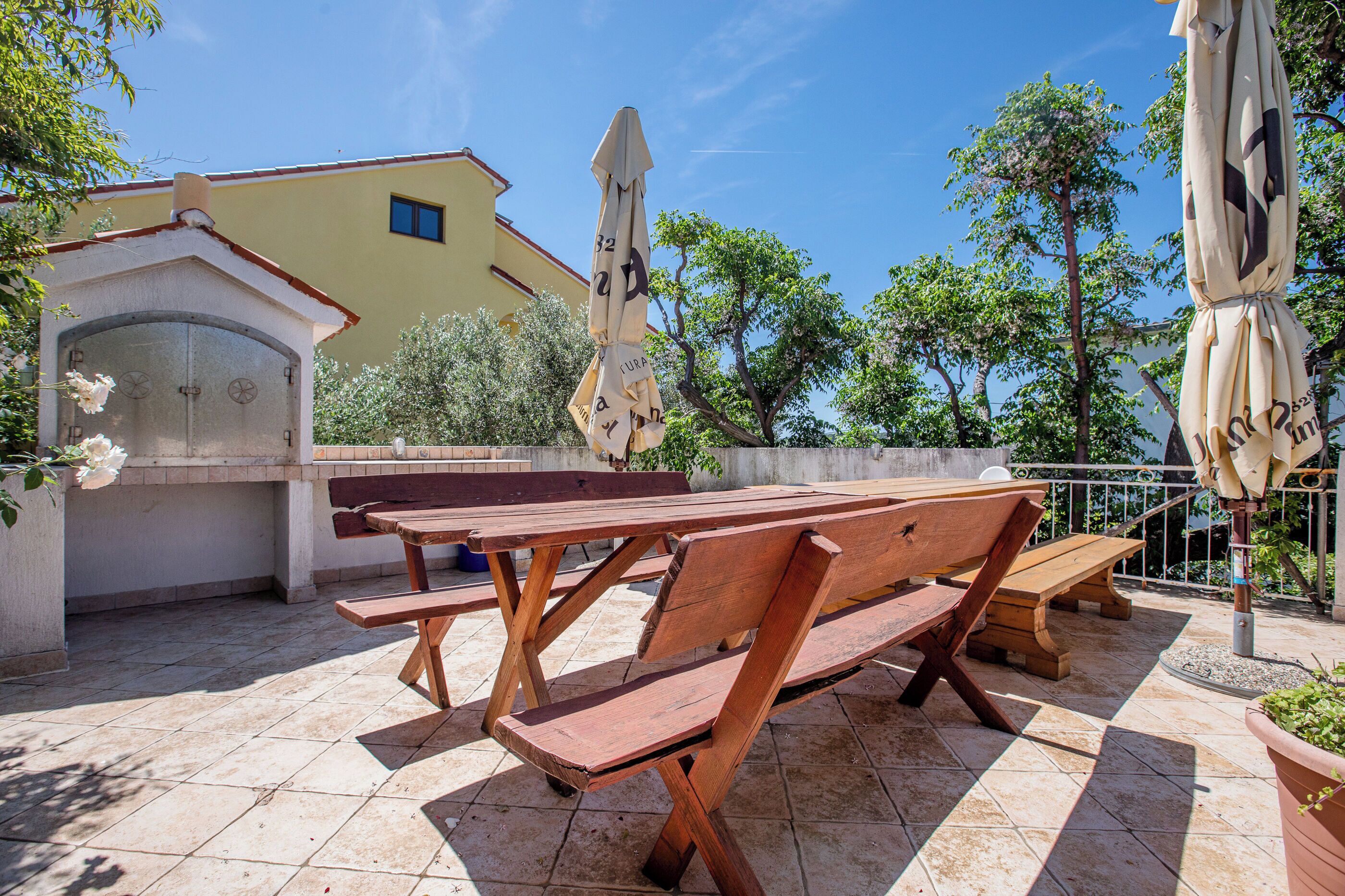 Un patio al aire libre con bancos de madera y sombrillas, un edificio de fachada amarilla y árboles al fondo.