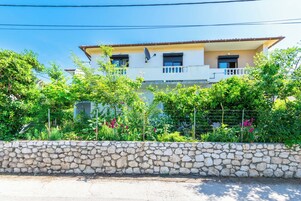 Una casa de dos pisos con un muro de piedra en el frente, rodeada de exuberante follaje y plantas con flores.