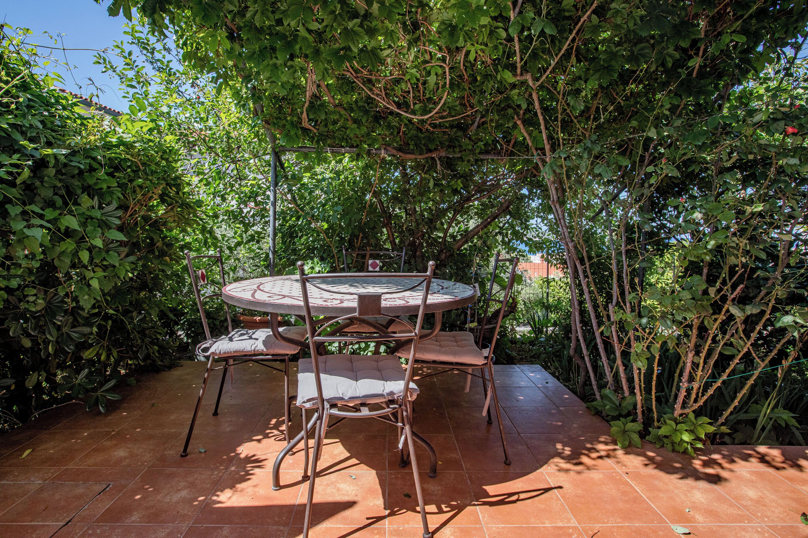 A round metal table with four chairs, surrounded by dense foliage and a tiled patio.