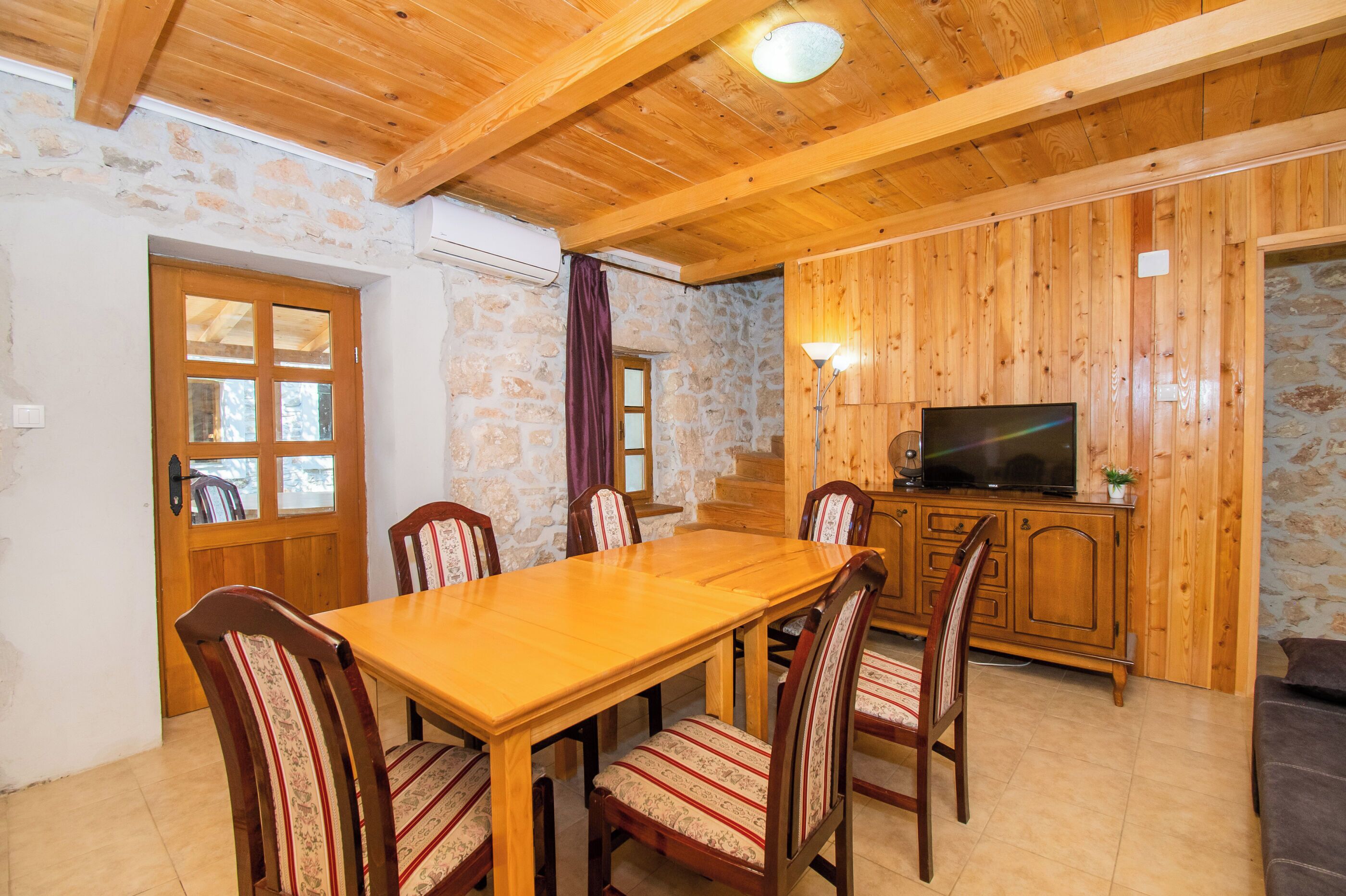 A wooden dining table with six chairs, a television on a wooden cabinet, and stone walls.
