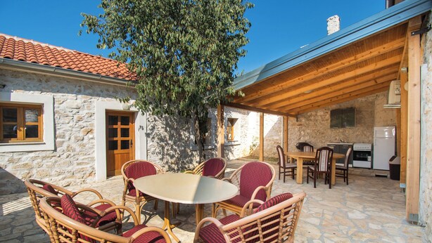 Un bâtiment en pierre avec un toit en tuiles, une pergola en bois, des meubles en osier et une table en pierre.