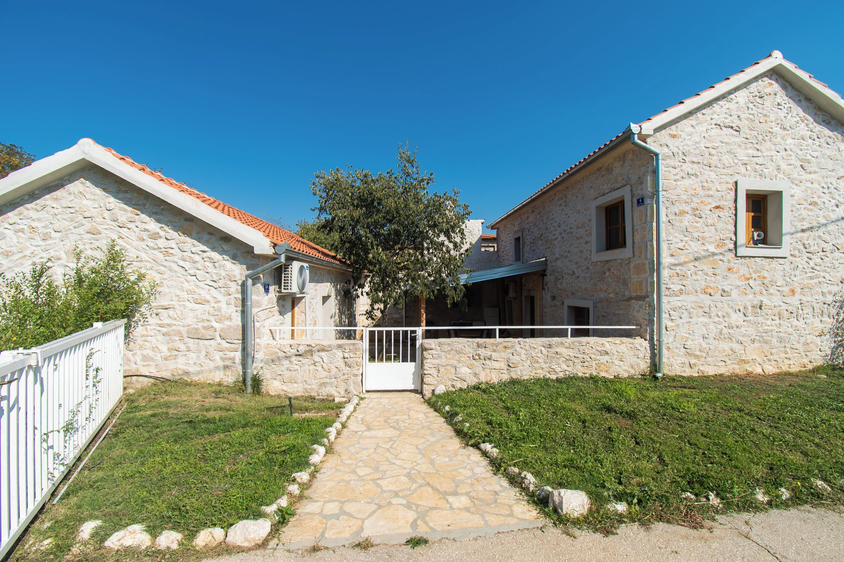 A stone house with a tiled roof, a paved entrance, and a white fence.