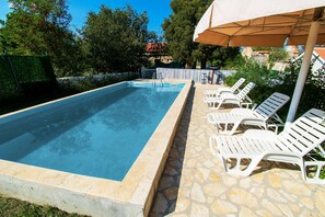 Ein rechteckiger Pool mit Steinbordurand, umgeben von weißen Liegestühlen und einem überdachten Bereich mit Sonnenschirm.