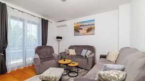Ein Wohnzimmer mit grauem Sofagarnitur, einem kleinen runden Tisch und einem großen Fenster mit transparenten Vorhängen.
