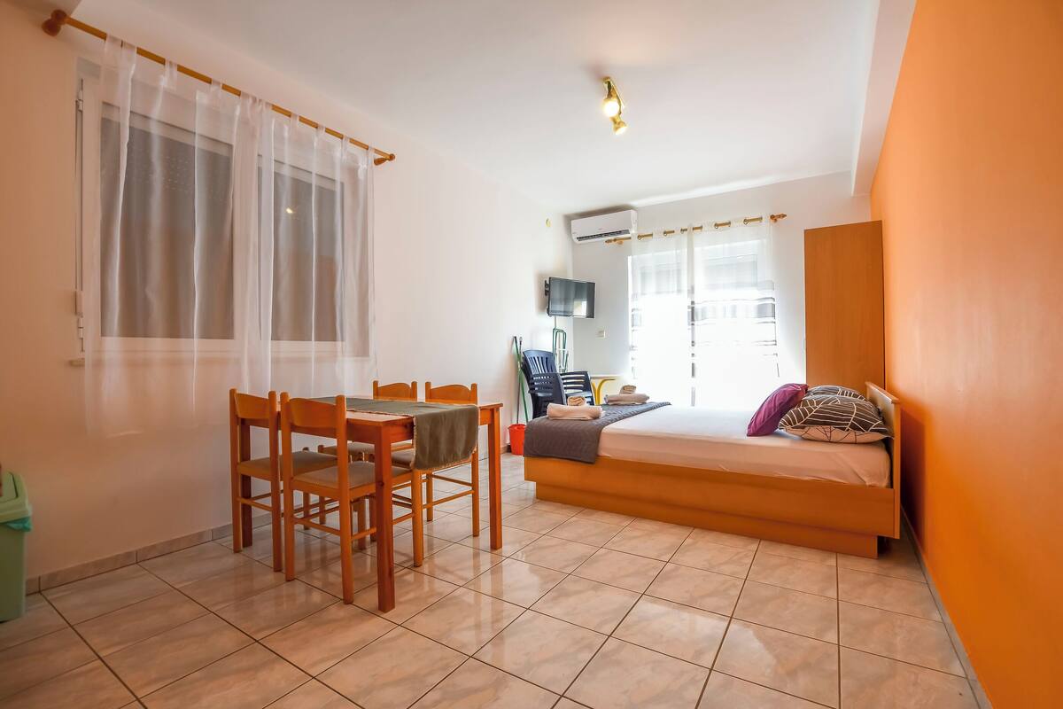 A compact room with a bed, a dining table with chairs, a TV, and an air conditioning unit.