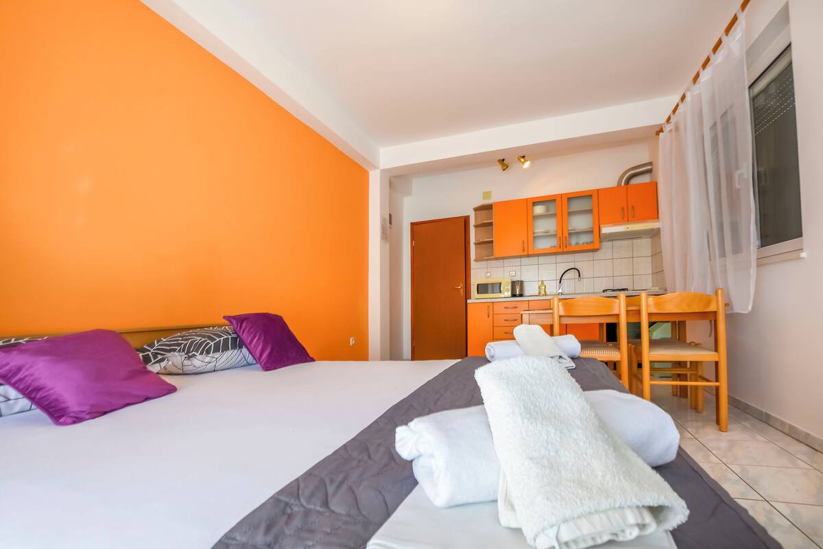 A hotel room with an orange wall, a bed with a grey and white bedspread, purple and white pillows, a white towel, a wooden chair, and a kitchenette with orange cabinets.