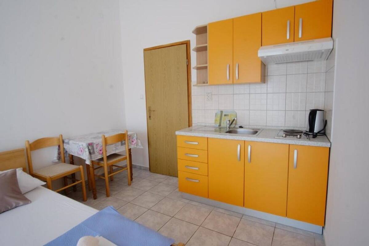 A small, compact room with a bed, a dining table with chairs, a kitchenette with yellow cabinets, and a wooden door.