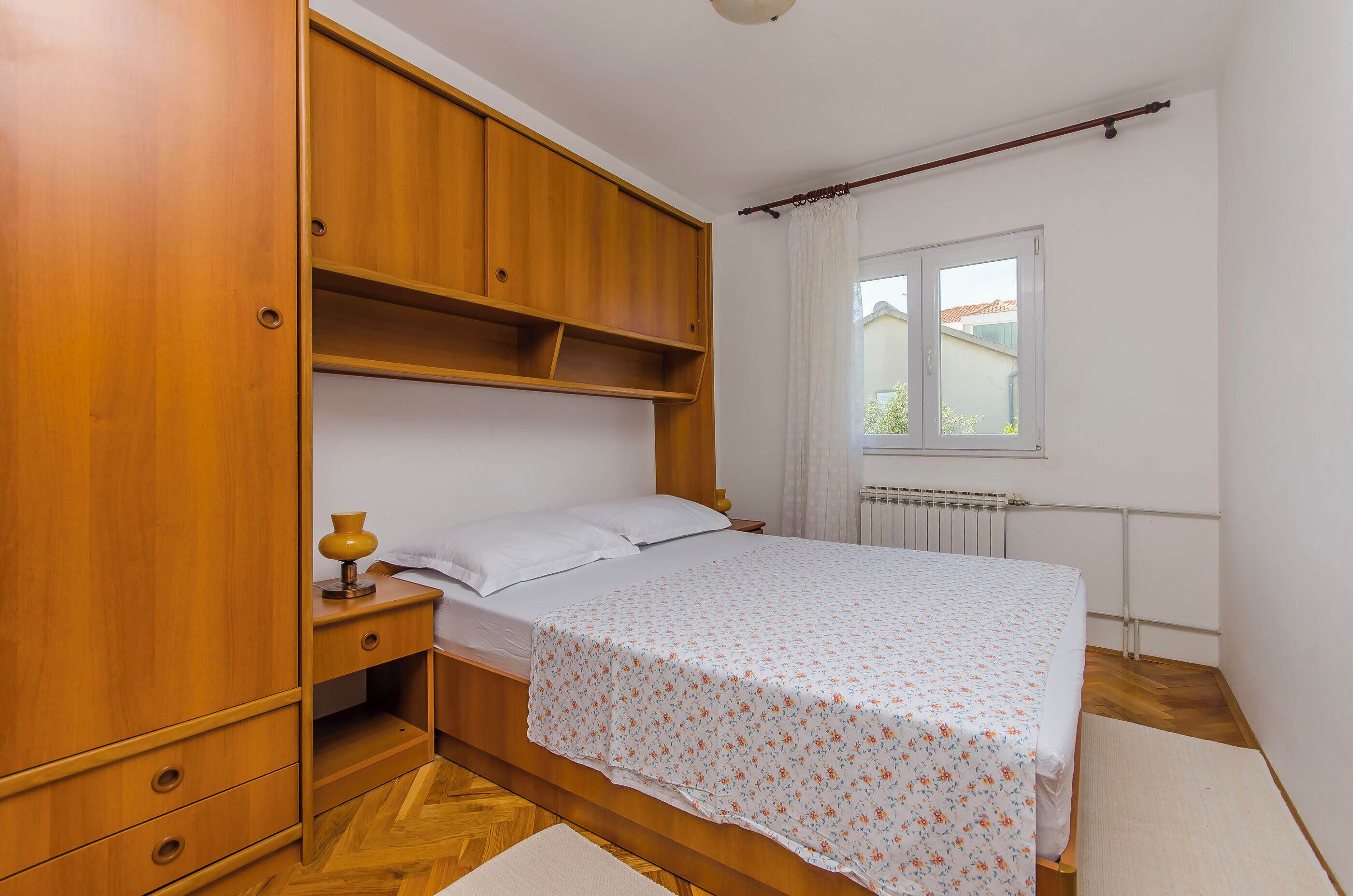 A bedroom with a wooden wardrobe, a bed with a floral bedspread, a nightstand with a lamp, and a window with curtains.