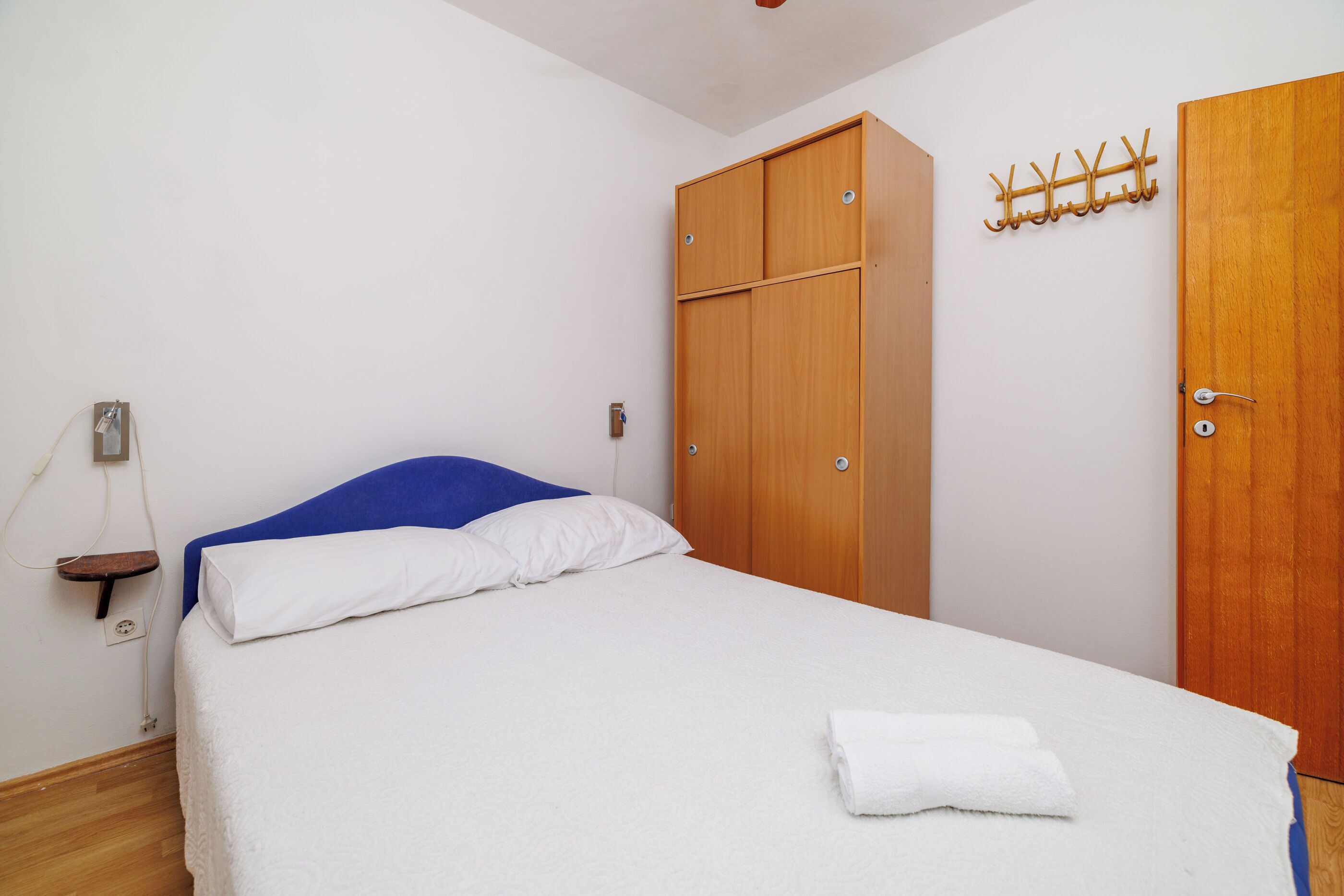 A neatly made bed with white bedding and a blue headboard, a wooden wardrobe with two doors, and a coat rack with hooks on the wall.