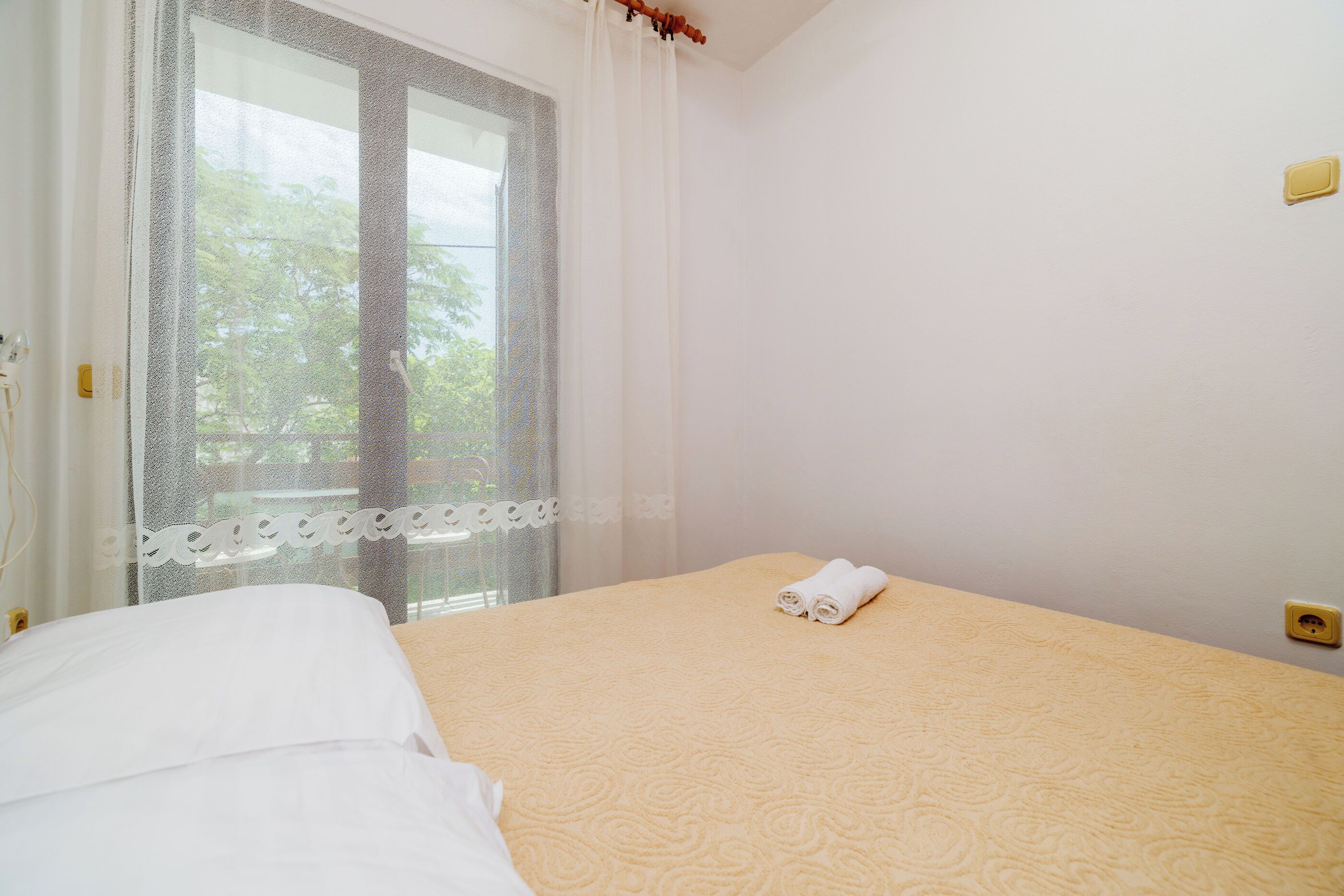 Een net opgemaakt bed met een beige sprei en witte lakens, een raam met dunne gordijnen, en een kleine gele schakelaar aan de muur.
