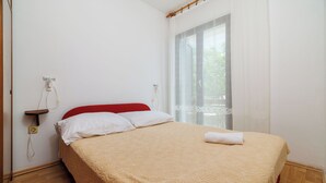 Een netjes opgemaakt bed met een rood hoofdeinde, witte kussens en een beige deken met dessin. Er staat een houten nachtkastje bij, met een lamp en een stopcontact op de muur.