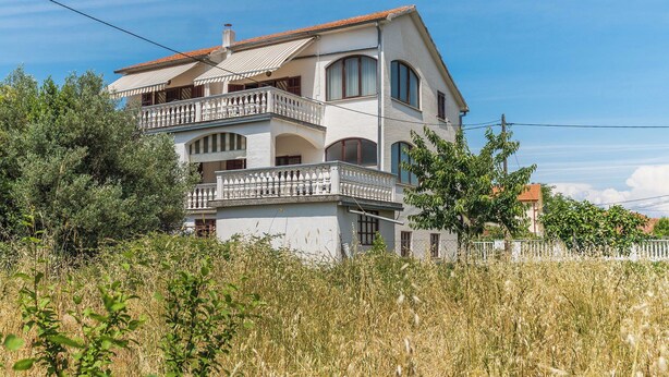 Une maison à deux étages avec un balcon, entourée d’herbes hautes et d’arbres.